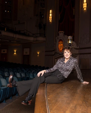 Woman in floral print shirt and black pants sitting on stage