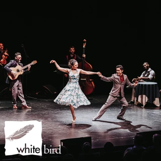 A woman in a floral dress and a man in a suit dance energetically on stage.