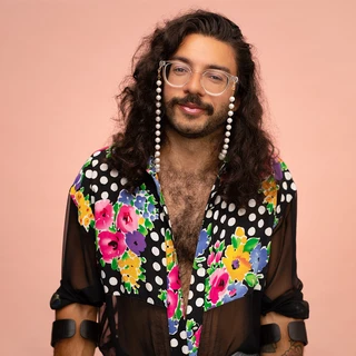 Smiling person (Alex Locust) with long curly hair and beaded glasses wears a sheer floral polka-dot shirt, posing confidently against a peach backdrop.