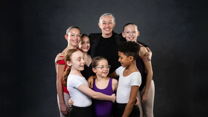 A joyful group of six young ballet students in colorful attire and their smiling teacher stand against a dark background, exuding warmth and camaraderie.