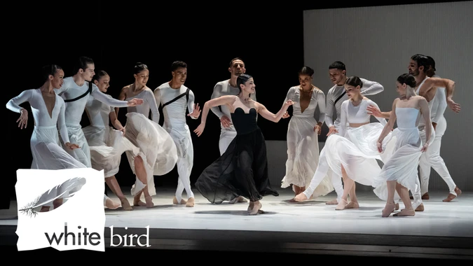 A group of dancers in elegant outfits, predominantly white with one in black, perform on stage with dynamic, graceful movements. The mood is energetic and dramatic. The "white bird" logo is in the bottom left corner.