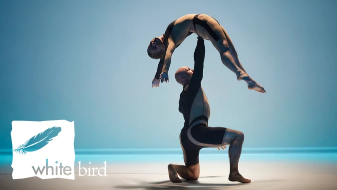 Two dancers perform an elegant lift against a soft blue background. One dancer kneels while gracefully holding the other aloft. 