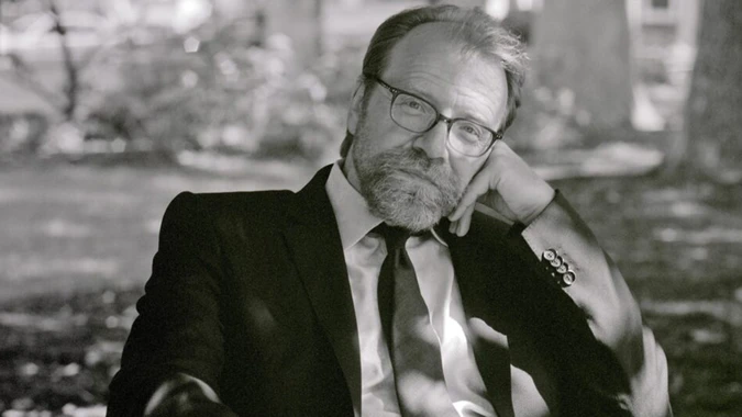 Black and white photo of George Saunders in glasses and a suit, sitting outdoors with a thoughtful expression. Sunlight filters through trees, creating dappled shadows.