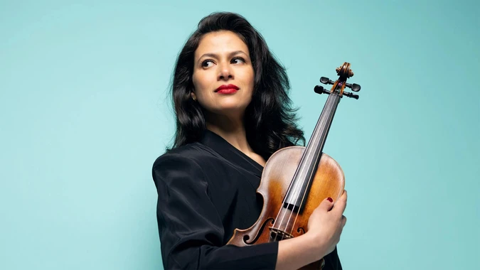 Photo of violinist Elena Urioste in black holding a polished violin against a turquoise background
