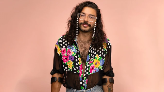Smiling person (Alex Locust) with long curly hair and beaded glasses wears a sheer floral polka-dot shirt, posing confidently against a peach backdrop.